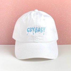 Pode incluir: Boné de beisebol branco com um texto bordado em azul "CRYBABY" com acentos de gotejamento.