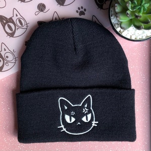 May include: Black beanie with a white embroidered cat face with angry eyes. The cat is facing forward.