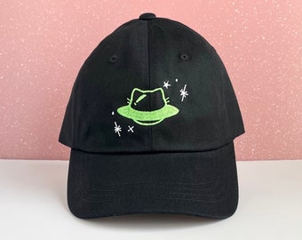 May include: Black baseball cap with a neon green embroidered design of a fedora-shaped UFO and white star accents. The cap has a curved brim and a button on top. A stylish accessory for casual wear.