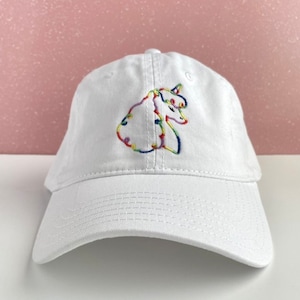 Puede incluir: Gorra de béisbol blanca con un diseño de unicornio bordado en arcoíris.