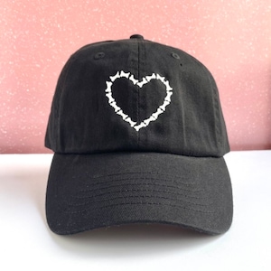 Könnte beinhalten: Eine schwarze Baseballkappe mit einem weißen gestickten Herzen aus kleinen Knochenformen.