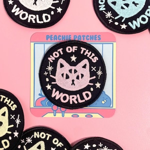 May include: Black embroidered patch featuring a pink cat face and the words "NOT OF THIS WORLD" in white. The patch is surrounded by stars and small asterisks. The background is pink.
