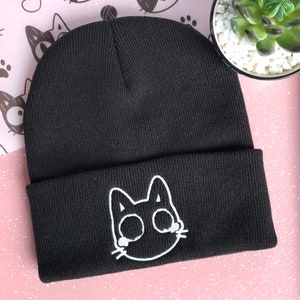 May include: Black knitted beanie with a white embroidered cat face on the folded brim. The cat has large eyes and whiskers. The beanie is displayed on a pink surface with a small succulent plant in a white pot.