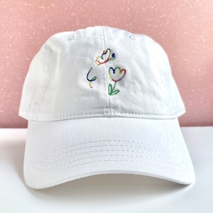 May include: White baseball cap with a colorful embroidered design of flowers and a swirl on the front.