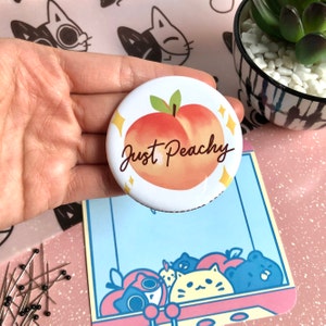 Peut inclure: Un bouton blanc avec une illustration de pêche et le texte "Just Peachy" en écriture brune. Le bouton a une bordure dorée pailletée.