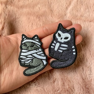 May include: Two embroidered iron-on patches featuring cats dressed up for Halloween. One patch is a black cat with a skeleton design and the other is a green cat wrapped in white bandages.
