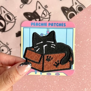 May include: Black cat embroidered patch with white eyes and nose, peeking out of a brown cardboard box with paw prints. The patch is on a white background with the text "Peachy Patches" in black.