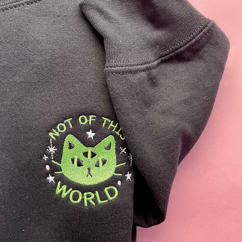 May include: Black sweatshirt with a green embroidered cat with multiple eyes and the text "Not of this world" in a circle around the cat.