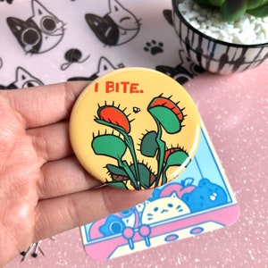 Puede incluir: Un botón amarillo con una ilustración de una planta de venus atrapamoscas verde y roja y el texto "I BITE."