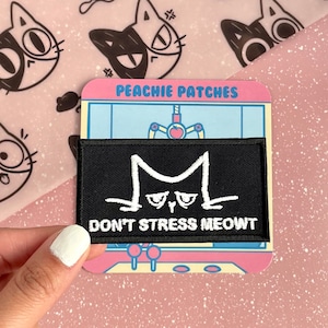 May include: Black iron-on patch with white embroidery of a cat face and the text "Don't Stress Meowt". The patch is on a pink background with a cartoon arcade game graphic that says "Peachy Patches".
