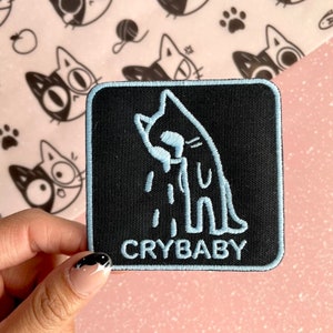 May include: A black iron-on patch with a blue outline of a crying cat and the text "CRYBABY" in blue.