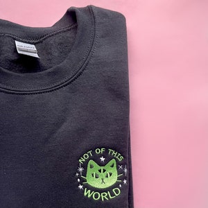 May include: Black sweatshirt with a green embroidered design of a cat with the text "Not of this world" in a circle.
