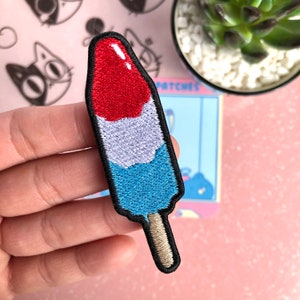 May include: A red, white, and blue embroidered popsicle patch with a brown stick. The patch is made of felt and has a stitched outline.