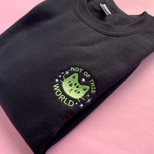 May include: Black sweatshirt with a green embroidered design of a cat with the text "Not of this world" around it.