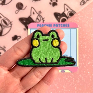 May include: Green embroidered frog patch with yellow eyes sitting on a green lily pad. The patch is on a hand and has the text "Peachy Patches" on a blue and white background.