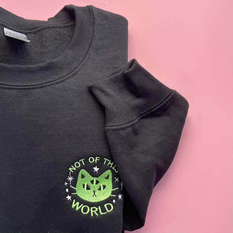 May include: Black sweatshirt with a green embroidered design of a cat with multiple eyes and the text "Not of this world".