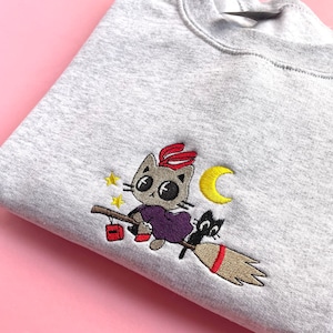 Puede incluir: Una sudadera gris con un diseño bordado de un gato montando una escoba con una luna creciente y estrellas en el fondo.