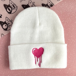 May include: White knit beanie with a pink dripping heart embroidered on the front.