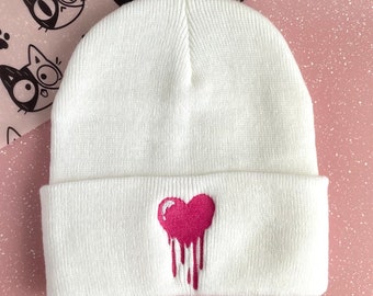 May include: White knitted beanie with a pink dripping heart embroidered on the front.