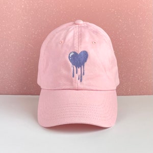 May include: A pink baseball cap with a purple dripping heart embroidered on the front.