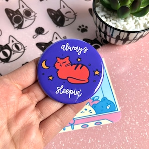 Könnte beinhalten: Ein lilafarbener Knopf mit einer roten Katze, die auf einem Halbmond mit Sternen schläft. Der Text auf dem Knopf lautet "always sleepin'"