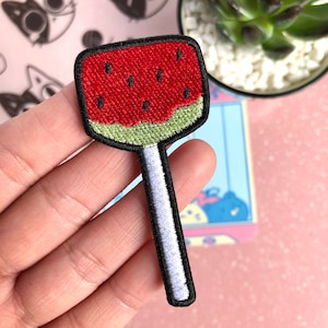 May include: A red and green watermelon lollipop patch. The watermelon is embroidered with black seeds and the stick is white with black outlines.