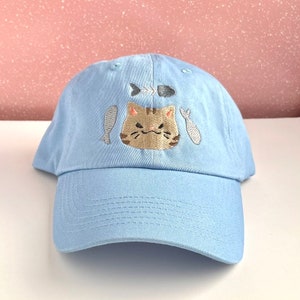 Peut inclure: Casquette de baseball bleu clair avec un visage de chat brodé et un motif d'arêtes de poisson. Le chat a une expression grincheuse. Deux poissons sont brodés de chaque côté du chat. La casquette est en tissu doux.
