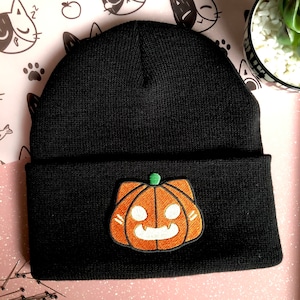 Könnte beinhalten: Schwarze Mütze mit einem Kürbis-Patch. Der Patch ist orange mit einem weißen Gesicht und einem grünen Stiel.
