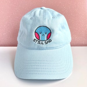 Könnte beinhalten: Eine hellblaue Baseballkappe mit einem gestickten Comic-Gesicht auf rosafarbenem Hintergrund und dem Text "STILL SAD".