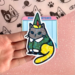 May include: A green and yellow glitter cat sticker with a pointy hat and a feather. The cat is wearing a green and gold patterned outfit. The sticker is on a pink background with white polka dots.