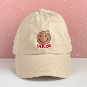 May include: A beige baseball cap with a curved brim. The cap features an embroidered design of a smiling cookie with the word "DULCE" in red below the cookie. The cap has a button on top.