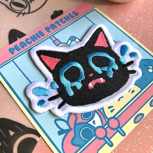 May include: A black cat with pink ears and blue teardrop-shaped eyes is embroidered on a white patch. The patch is surrounded by a white border and has the text "Peachy Patches" in pink letters on a blue background.
