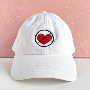 May include: White baseball cap with a curved brim. The cap features an embroidered design of a red heart with a black line through it, inside a black circle. The hat is made of a soft, durable fabric.