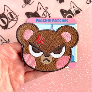 Puede incluir: Parche bordado con un oso pardo enfadado con mejillas rosas y un símbolo rojo de enfado. El parche es de "Peachie Patches". El fondo tiene un patrón de gatos rosas.