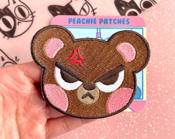 Puede incluir: Parche bordado con un oso pardo enfadado con mejillas rosas y un símbolo rojo de enfado. El parche es de "Peachie Patches". El fondo tiene un patrón de gatos rosas.
