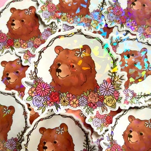 May include: A stack of holographic stickers featuring a brown bear with a flower crown. The bear is surrounded by a wreath of colorful flowers. The stickers are perfect for adding a touch of whimsy to your laptop, water bottle, or planner.