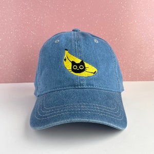 May include: Blue denim baseball cap featuring an embroidered design of a black cat inside a yellow banana. The cap has a curved brim and an adjustable strap at the back. The hat is a casual accessory.