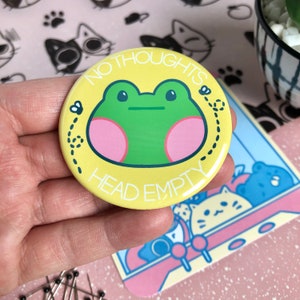 May include: A yellow button with a green frog and the text "NO THOUGHTS HEAD EMPTY" in black. The frog has pink cheeks and there are two black flies with white wings on either side of the frog.