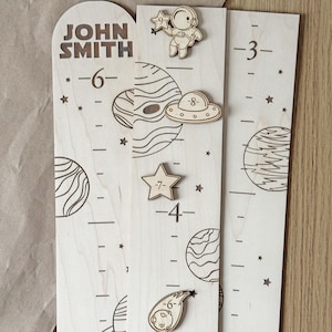 May include: A wooden growth chart with a space theme. It features planets, stars, a rocket, a UFO, and an astronaut. The chart is personalized with the name "JOHN SMITH".