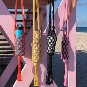 May include: Four macrame water bottle holders in orange, yellow, black, and pink, each with a matching tassel. The holders are suspended from a pink lifeguard stand on a sunny beach. A blue water bottle is in the orange holder.