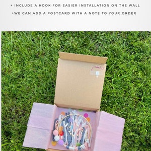 May include: A cardboard box with a pink tissue paper lining, containing a handmade dream catcher with white, pink, and blue ribbons, wooden beads, and colorful felt circles. The box is labeled with the text "Beautiful Package for Secure Transportation" and includes a hook for easy wall installation.