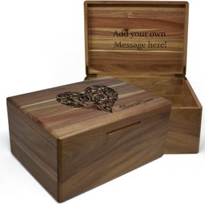 May include: A wooden box with a heart-shaped design engraved on the lid. The box is made of walnut wood and has a natural finish. The lid is open and the inside of the box is visible. The text "Add your own Message here!" is engraved on the inside of the lid. The text "Blessed and Grace" is engraved on the bottom of the box.