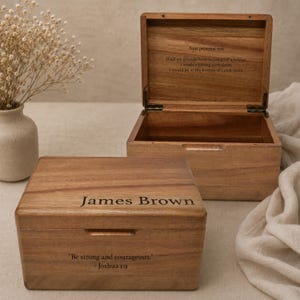 May include: Two rectangular wooden boxes with a natural grain finish. One box is closed and engraved with the name James Brown and the text Be strong and courageous. - Joshua 1:9. The other box is open, revealing an interior engraved with text.