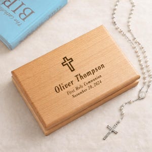 May include: A light-colored wooden box with a cross symbol and the name "Oliver Thompson" engraved on the lid, along with the text "First Holy Communion November 20, 2024." A rosary with a silver cross is next to the box.