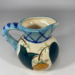 May include: A small, blue and white ceramic pitcher with a pineapple design. The pitcher has a round body and a handle.