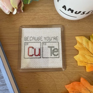 May include: Square coaster with a cross-stitched design. The coaster features the phrase "BECAUSE YOU'RE" above two boxes with "Cu" and "Te" inside, forming the word "cute". The coaster is in a clear plastic holder.