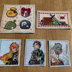 May include: Five embroidered cross-stitch patterns on white fabric with black stitching. The patterns include a green leaf, a blue and white circle, a red and white gift box, a yellow bag, and a red chest with a sword, a green frog, and a book.
