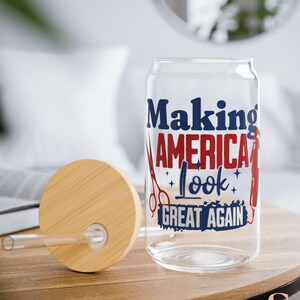 May include: A clear glass can-shaped tumbler with a wooden lid and straw. The tumbler features the text "Making America Look Great Again" in red and blue, along with barber tools. The lid is made of light brown wood.