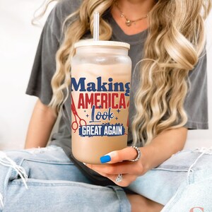 May include: A clear glass tumbler with a bamboo lid and straw, filled with a light brown beverage. The tumbler features the text "Making AMERICA Look GREAT AGAIN" in red, white, and blue, with scissors and star graphics.