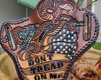 May include: A brown leather holster featuring a cobra design, a gun, and an American flag. The text "DON'T TREAD ON ME" is visible. The holster is crafted with detailed tooling and stitching, showcasing a unique design.